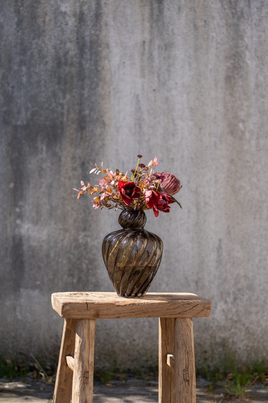 Dekorativ høstbukett med kunstige blomster og greiner i en mørk vase på en rustikk trekrakk.