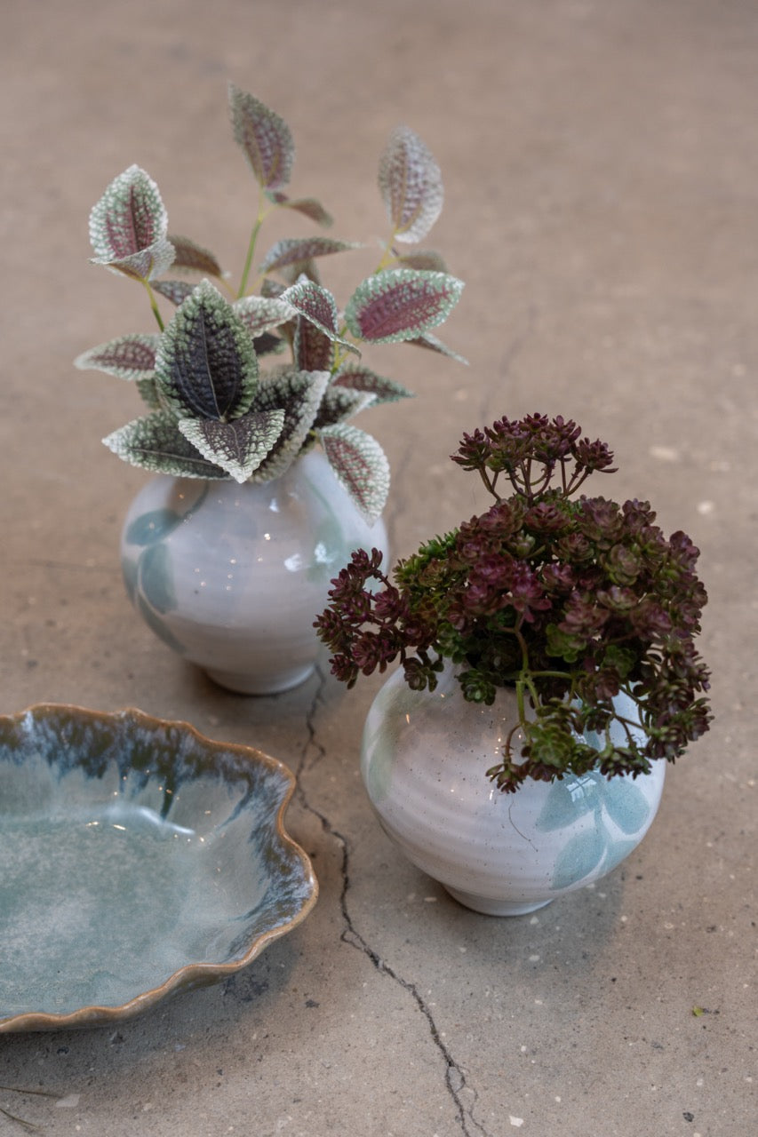 Kunstig grein med flekkete grønne og rosa blader i en liten keramikkvase sammen med mørke høstblomster.