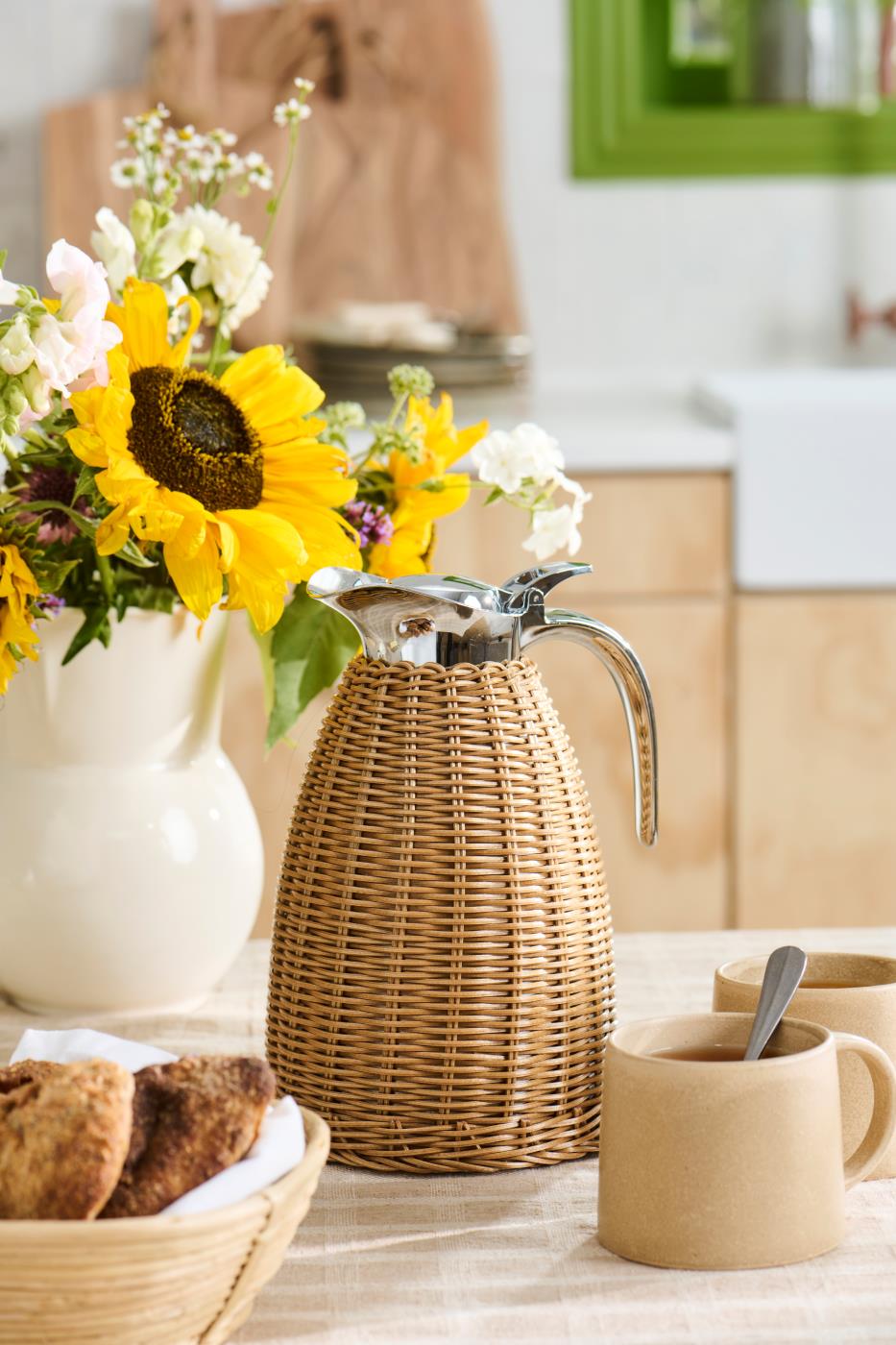 Termoskanne i brun polyrattan på dekket kaffebord med solsikker, keramikk og nybakt brød.
