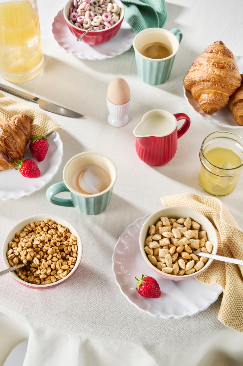 Melkekanne i jordbær- og myntefarge på frokostbordet med croissanter, müsli, kaffe og fersk frukt