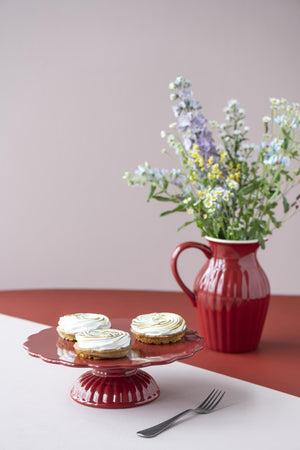 Rød myntekanne med blomster på sommerbord med kakefat og lyserosa bakgrunn