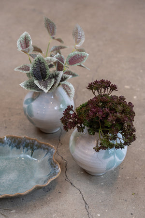 Kunstig grein med flekkete grønne og rosa blader i en liten keramikkvase sammen med mørke høstblomster.