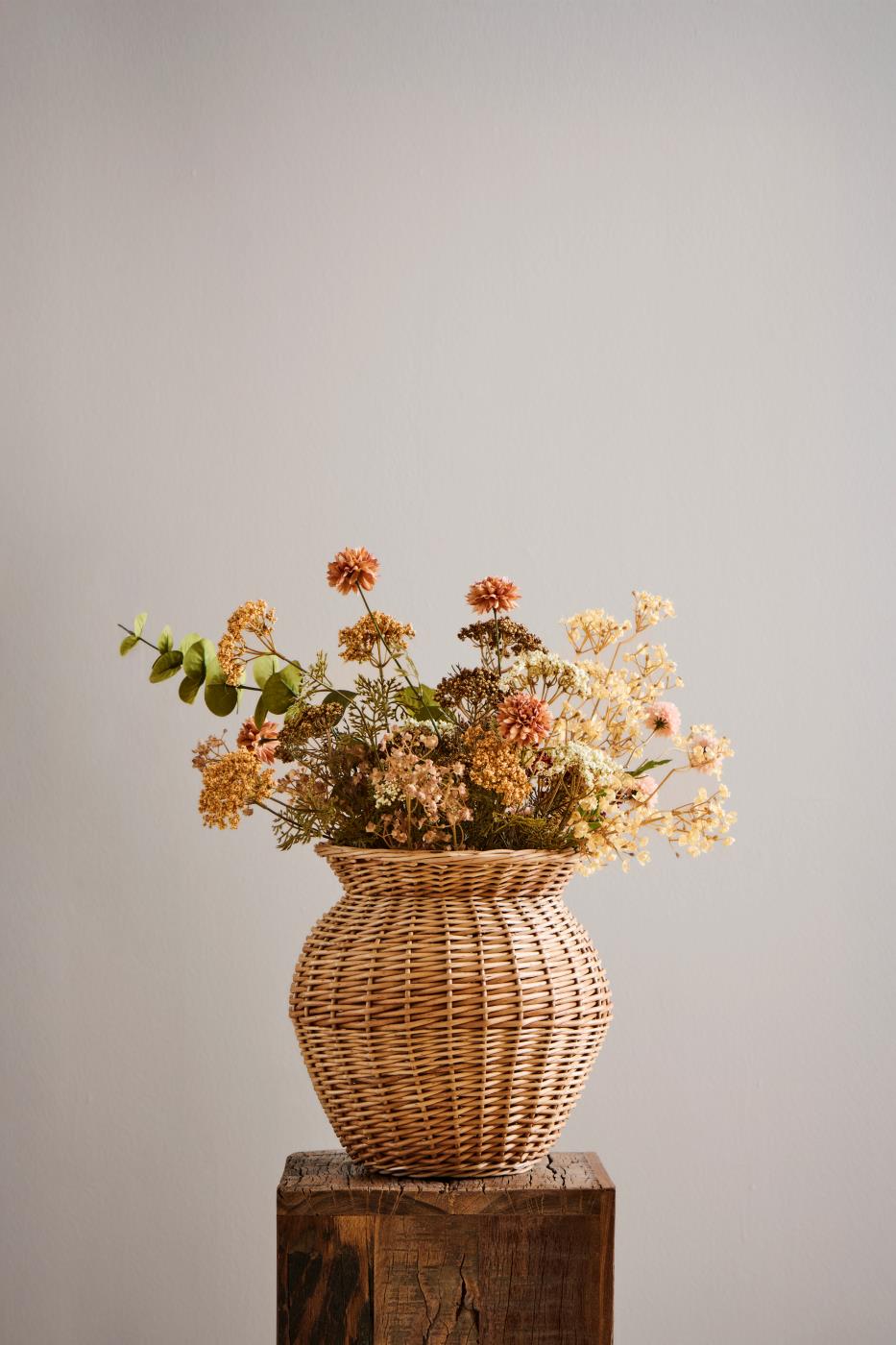 Høstbukett med kunstige blomster i hvite, oransje og gylne nyanser i en rustikk flettet vase.